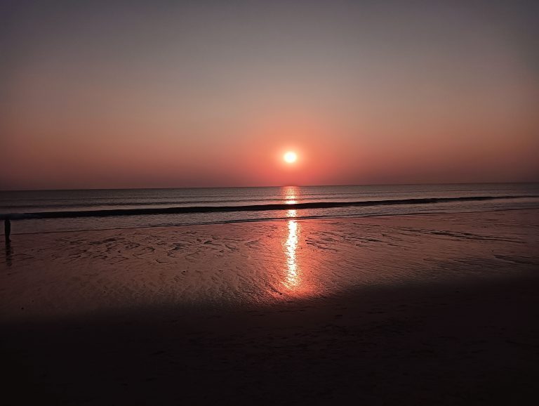 A serene beach sunset with the sun low on the horizon, casting a warm orange glow over the calm ocean and wet sand reflecting the sunlight.