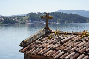 A rustic stone roof with a cross on top, overlooking a calm body of water with a shoreline in the background. The roof has patches of moss and small plants growing between the tiles. In the distance, there are hills and a few scattered buildings surrounded by lush greenery.