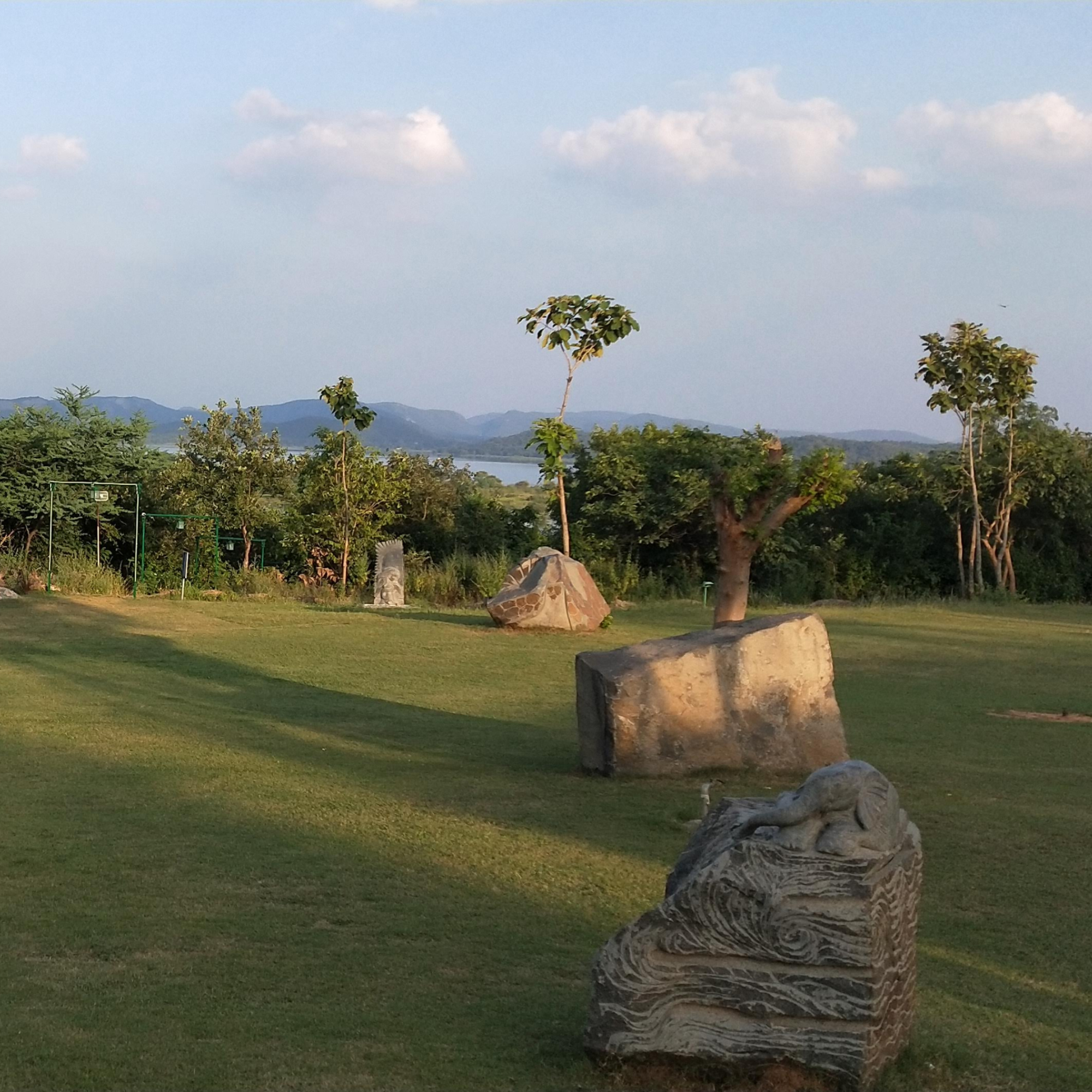 A scenic outdoor landscape featuring a grassy field with several large stones and small trees scattered throughout. In the background, there is a view of distant hills and a body of water, under a partly cloudy blue sky.