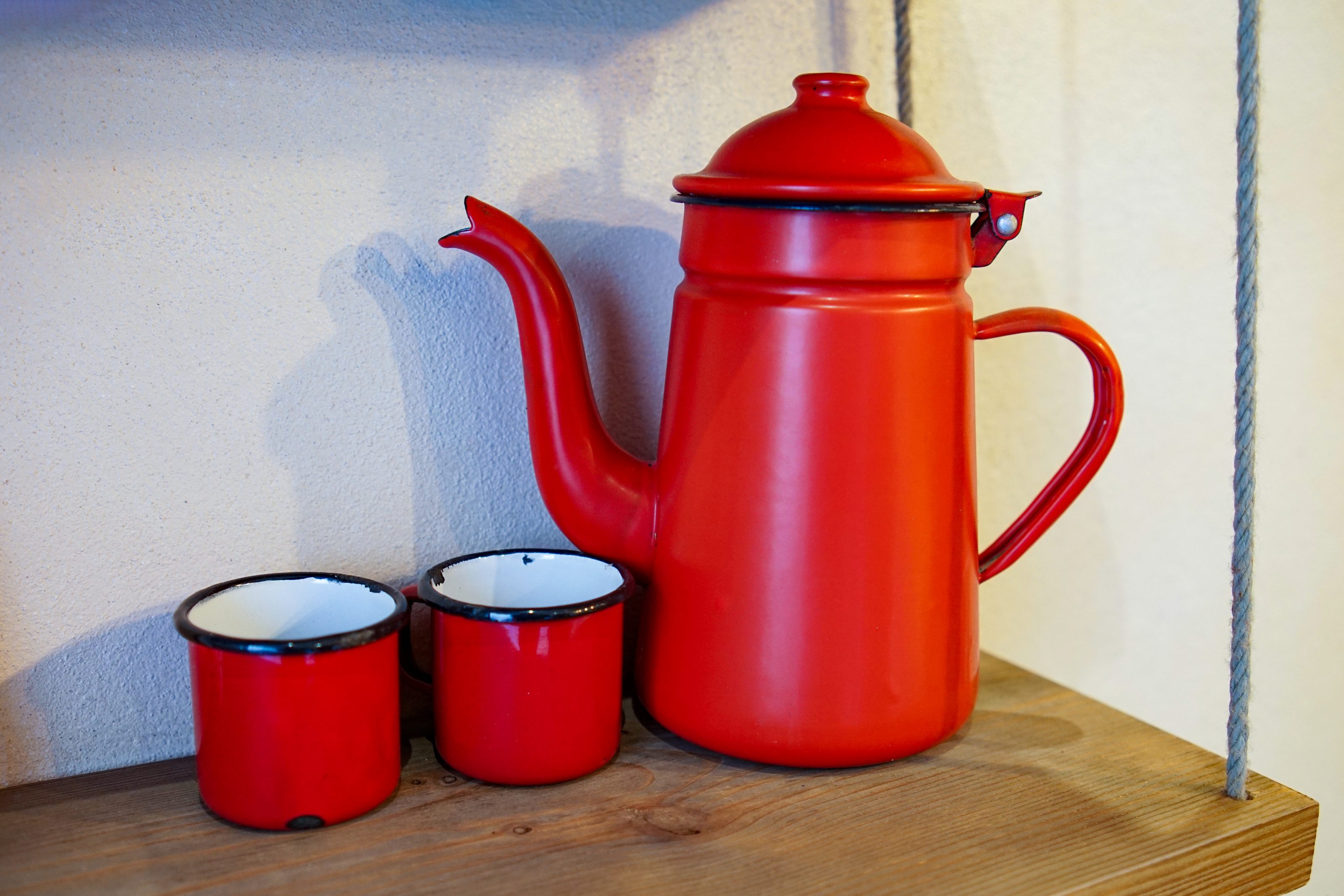 A red enamel kettle with a lid and spout, accompanied by two matching red enamel mugs with white interiors, placed on a wooden shelf.