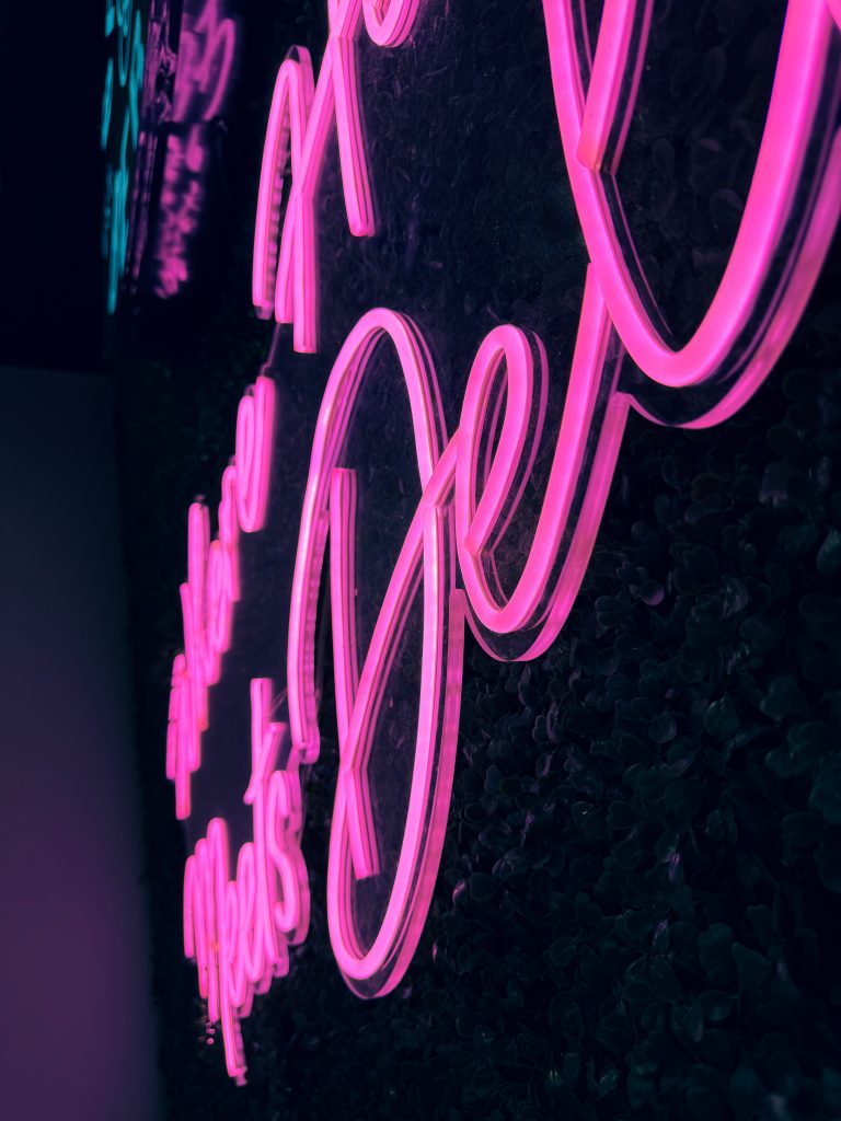 A close-up of a neon sign featuring pink cursive letters against a dark, textured background. The sign glows brightly, casting light onto the surrounding surface.