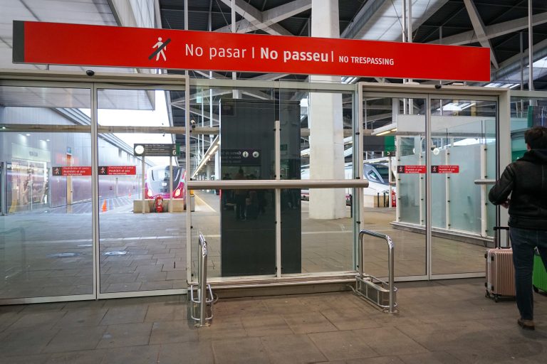 A glass door at a train station with a red “No Trespassing” sign in multiple languages. There’s a person with a suitcase nearby, and a train is visible on the other side of the door.