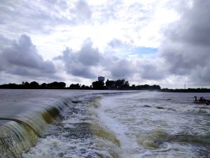 A wide-angle view of water cascading over a low dam, creating a series of small waterfalls. The sky above is cloudy with patches of blue, and there is a silhouette of trees and a building in the distance. 