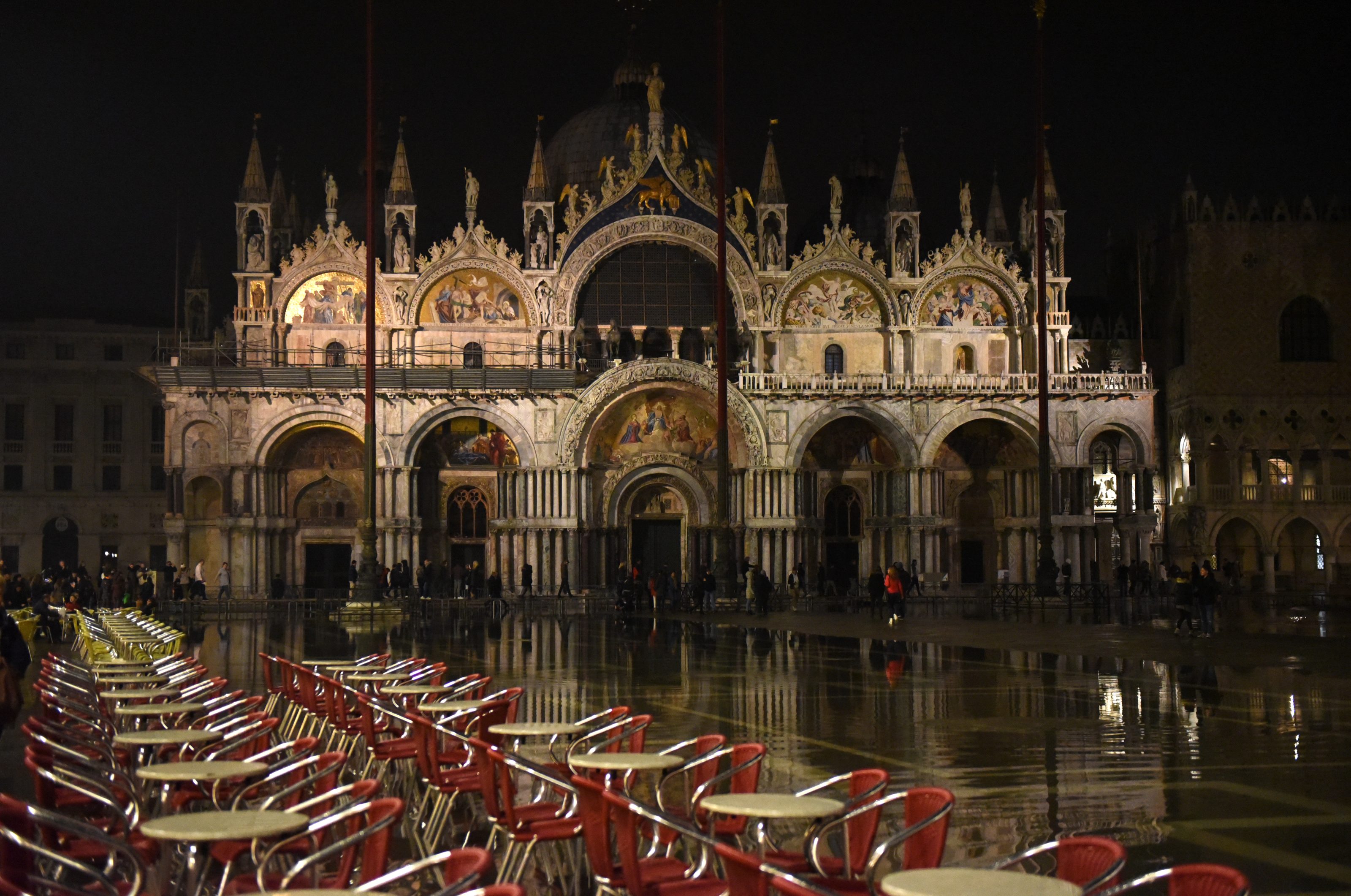 Venice's St. Mark's Square and Basilica at night with a row of chairs and tables on a café terrace in front. The ground is wet from the September spring tides.