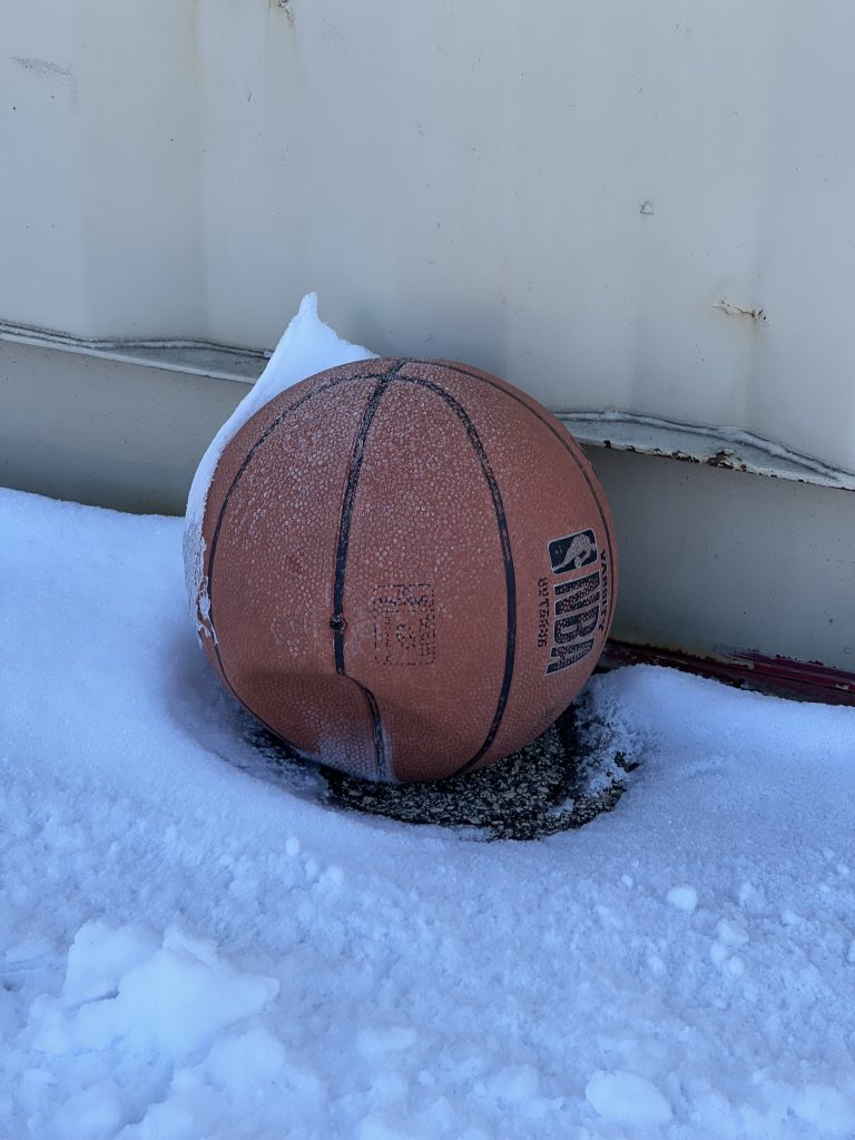 A slightly deflated basketball sitting next to a metal wall and on top of a frosted snow ground covering.