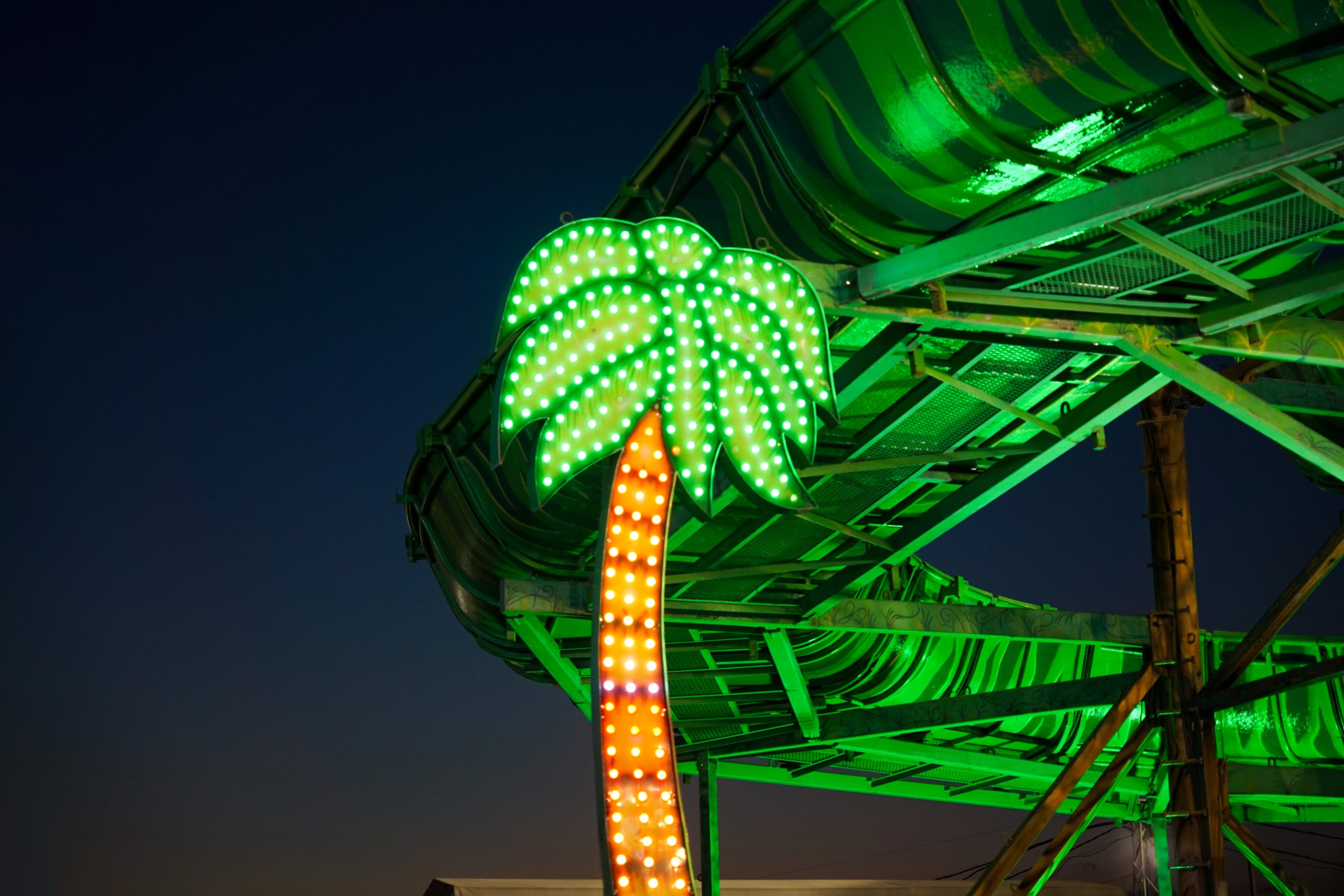 Illuminated palm tree sign in front of a swirling green amusement park slide, with a dark evening sky in the background.