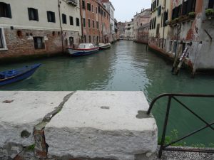 A picturesque canal in Venice, Italy, lined with weathered buildings featuring brick and stucco facades. Several small boats are moored along the calm, greenish water. There's a rustic stone wall with a metal railing in the foreground.