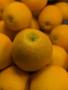 Close-up of bright oranges piled together, with one in focus showing a textured peel and green stem. Background oranges are blurred, creating depth. Soft lighting highlights colors, texture, and subtle shadows.