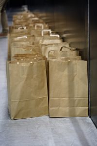 A row of brown paper bags, with handles haphazardly akimbo, lined up against a wall on a concrete floor.