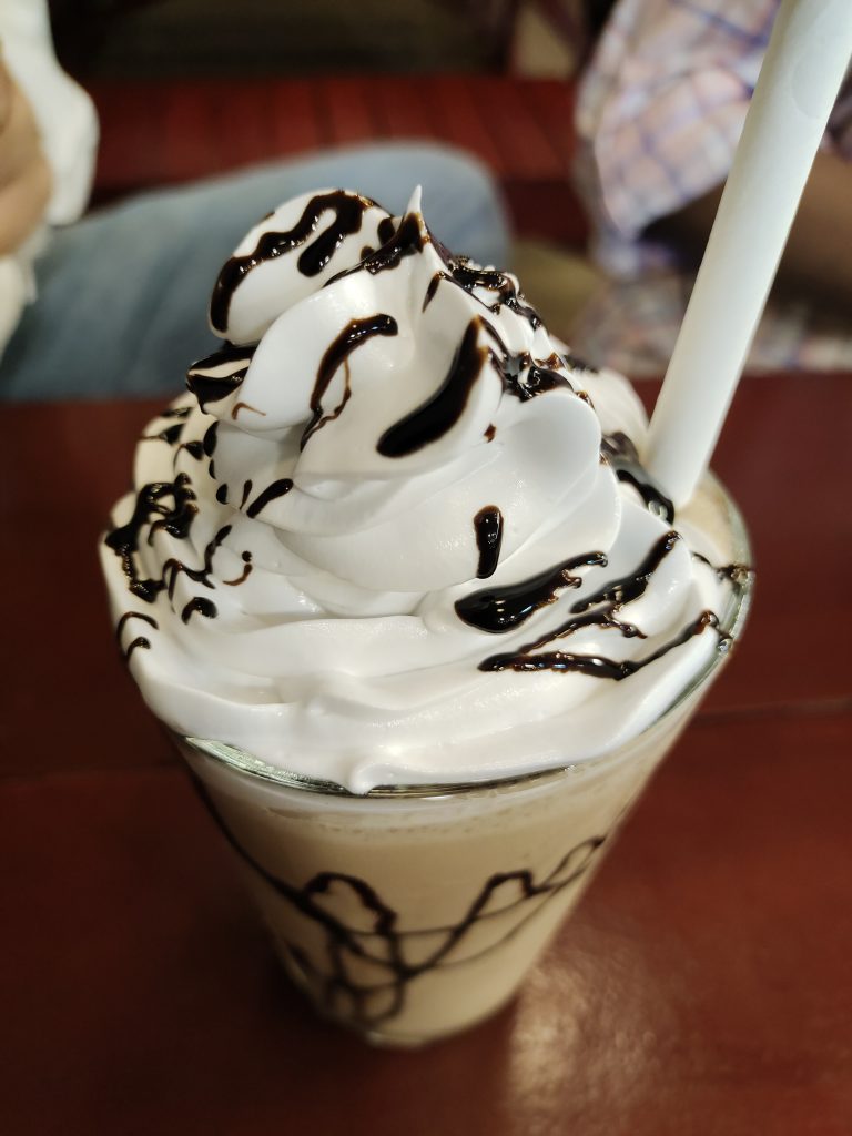 A glass filled with a creamy, light brown beverage topped with a swirl of whipped cream drizzled with chocolate syrup and a white straw.