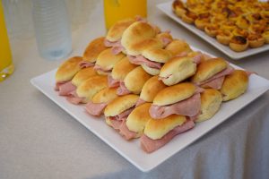 A platter of small sandwiches with thinly sliced meat inside soft rolls, arranged in a pyramid on a white plate. In the background, there are trays of small pastries and clear bottles filled with orange-colored drinks.