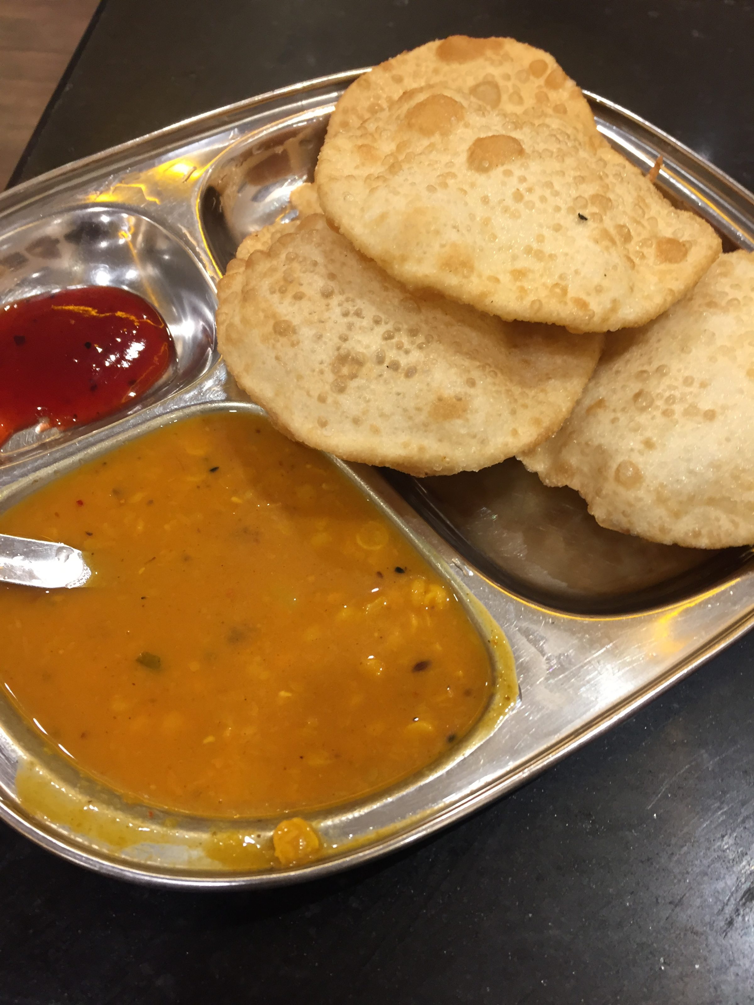 A stainless steel plate with three sections containing Indian food: a stack of puris, a serving of red chutney or sauce, and a portion of yellow dal or curry with a spoon.