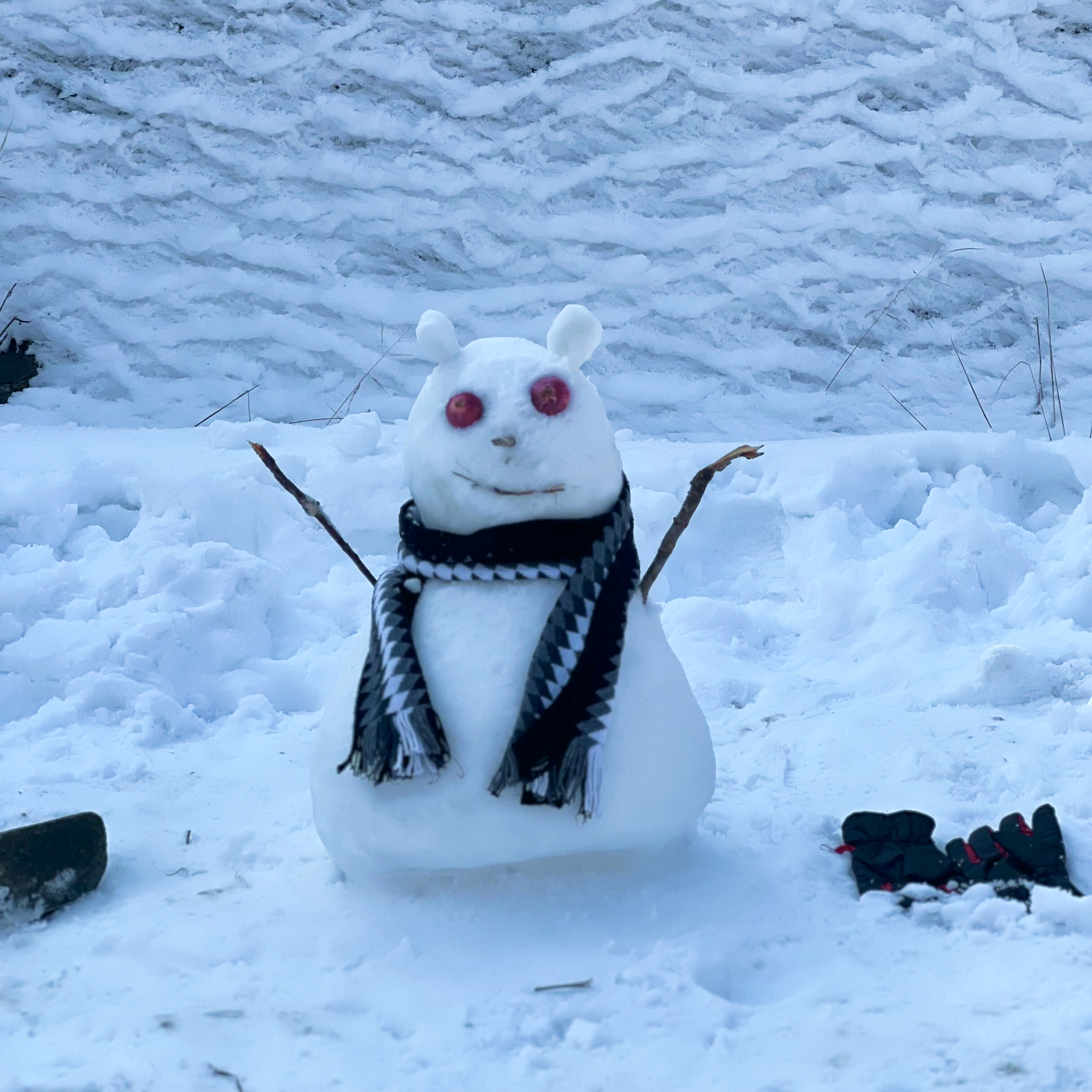 A small snowman with red eyes, a scarf, and stick arms, sitting in a snowy landscape.