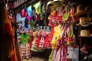 A vibrant market stall displaying colorful Flamenco dresses with polka dots hanging on racks.  Additionally, there are shelves with shirts in various colors and designs and a row of straw hats hanging on the right side.