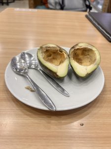 A sliced avocado with its pit removed, placed on a white plate alongside two metal spoons, set on a wooden table.