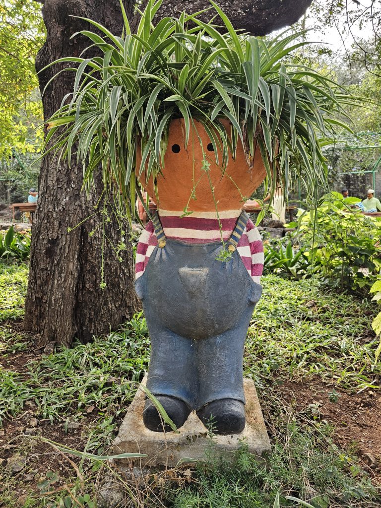 A whimsical garden sculpture of a figure with a flower pot as its head. The pot is filled with spider plants, creating the appearance of wild green hair.