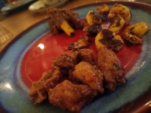 A close-up of a colorful plate with a variety of grilled and fried meats, including shrimp and possibly chicken, arranged in sections. The food is seasoned and appears to be well-cooked, with some charring visible. The plate has a red center with a blue rim.