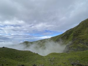 A scenic view of green hills partially covered in mist, under a sky filled with clouds. The landscape features rolling hills and rocky outcrops, creating a serene and expansive natural vista.