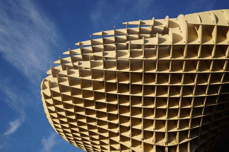 A close-up view Metropol Parasol, World’s largest wooden structure. A huge, intricate wooden lattice structure with a geometric pattern against a clear blue sky with scattered white clouds.