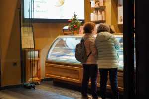 Two people in winter jackets are standing in front of a gelato display case inside an ice cream shop. A small decorated Christmas tree is visible on the counter, and shelves with cups are in the background.