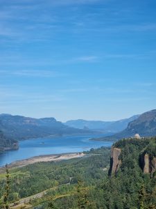 A view of the blue Columbia River meandering through a lush, tree-covered valley set against a clear blue sky.
