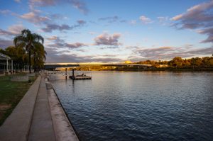 A waterfront scene at sunset with a calm river in the foreground, a bridge in the middle distance, and a tree-lined shoreline. The sky is filled with clouds reflecting the warm colors of the setting sun. A small pier extends into the river from the left, bordered by a pathway with palm trees and grass