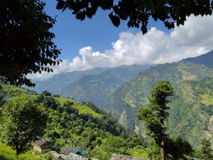 A peaceful hillside village surrounded by green fields and trees. The fields are neatly arranged in steps, going down the hill. Small houses with simple roofs are scattered across the area. In the background, tall mountains rise high, partly covered by white clouds in the blue sky. The scene is framed by tree branches at the top, adding a cozy feel