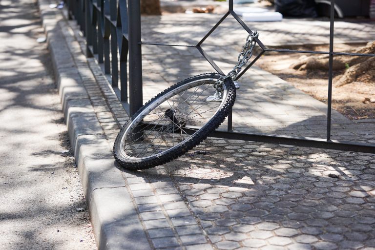 A bicycle wheel chained to a metal fence, with the rest of the bicycle missing. The wheel is resting on a cobblestone walkway, in a partially shaded area.