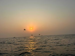 A serene ocean view at sunset with the sun low on the horizon, casting a warm glow over the water. Several parasailers are visible in the distance, and a couple of boats are barely discernible on the ocean's surface.