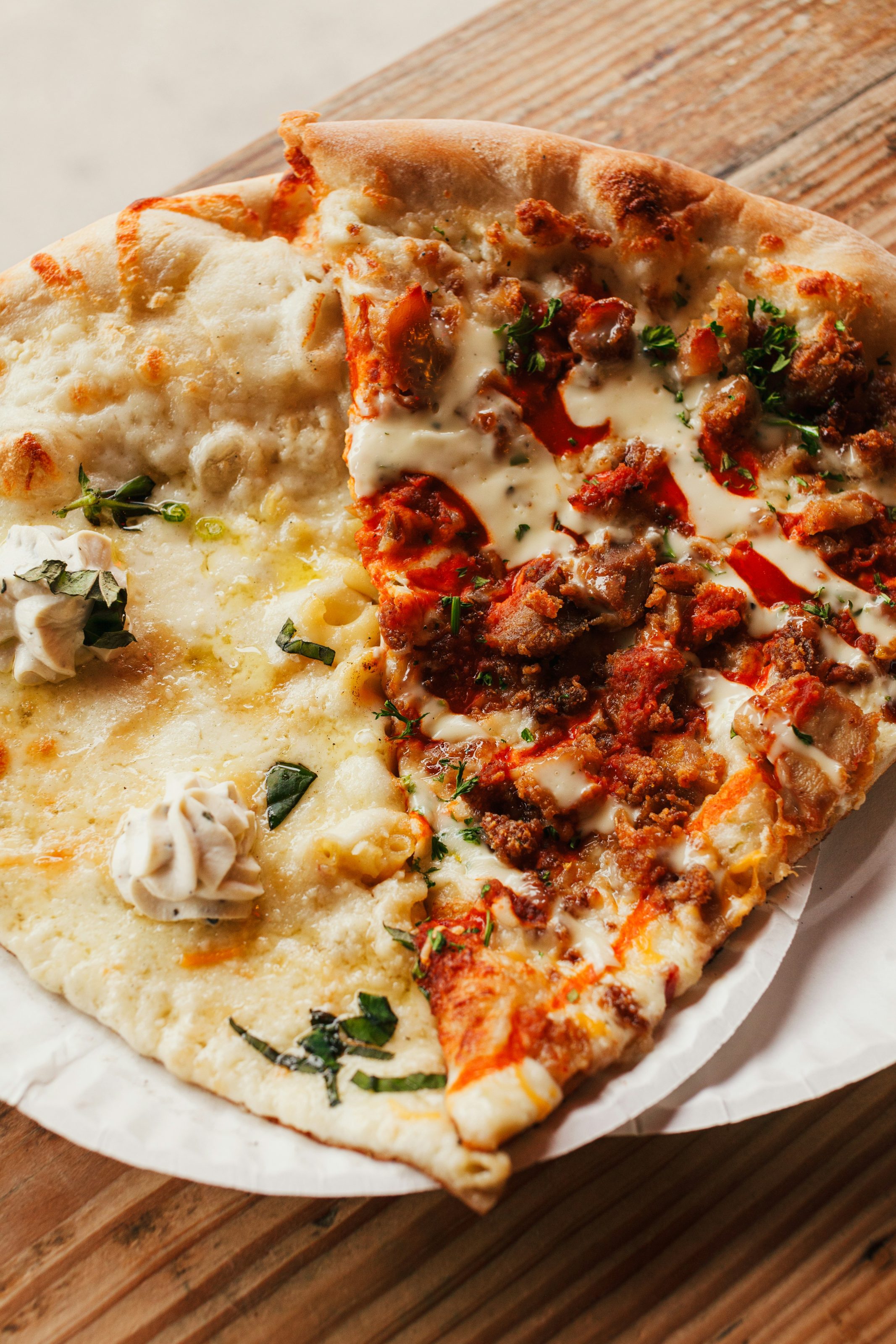A close-up of two slices of pizza on a paper plate sitting on a wooden table. The slice on the left is topped with creamy sauce, herbs, and cheese. The slice on the right features meat, red sauce, and a drizzle of creamy dressing.