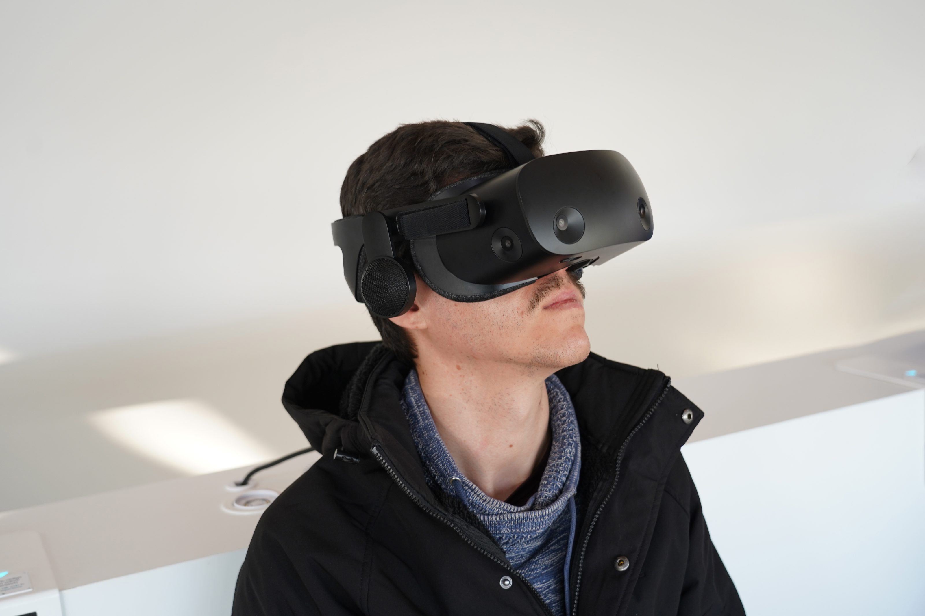 A person wearing a black virtual reality headset, looking slightly upward. They are dressed in a dark jacket and a blue sweater, standing indoors with a plain white background.