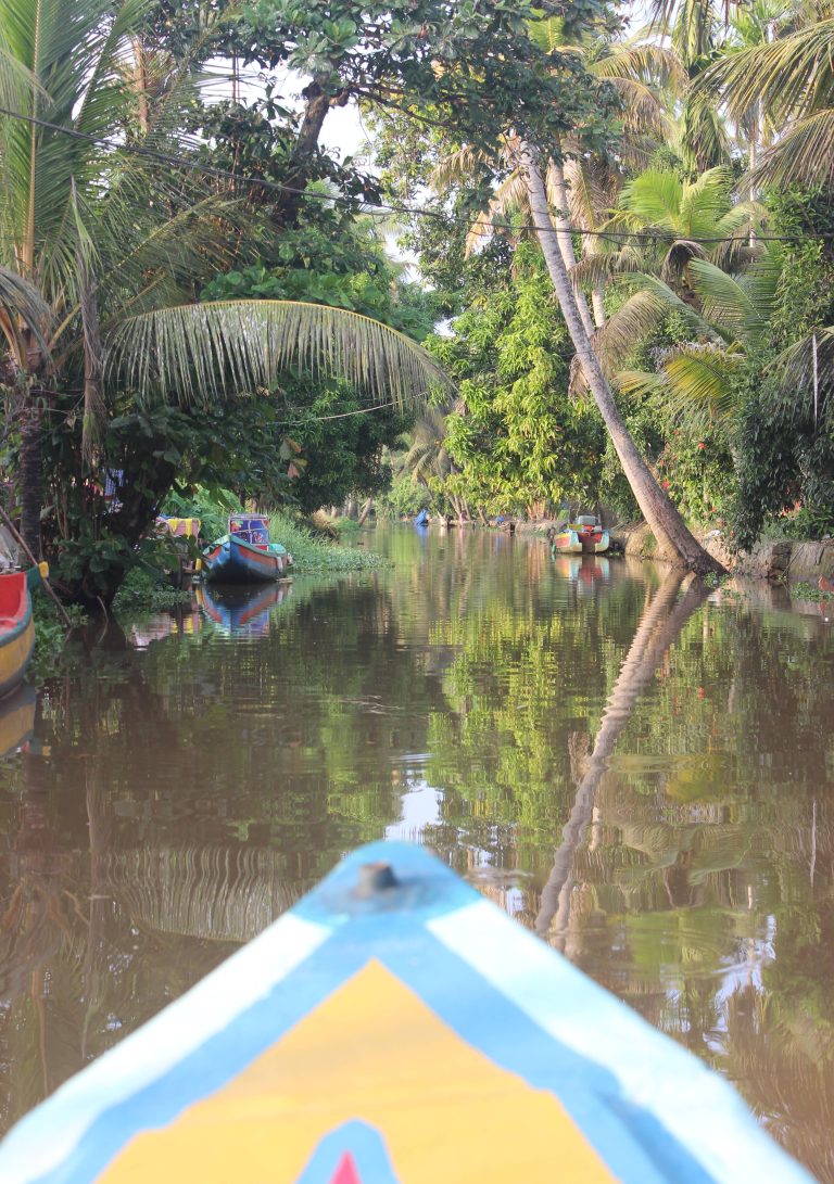 View from the bow of a colorful boat navigating a narrow, calm river surrounded by lush green trees and vegetation. Several other boats are moored along the riverbanks.