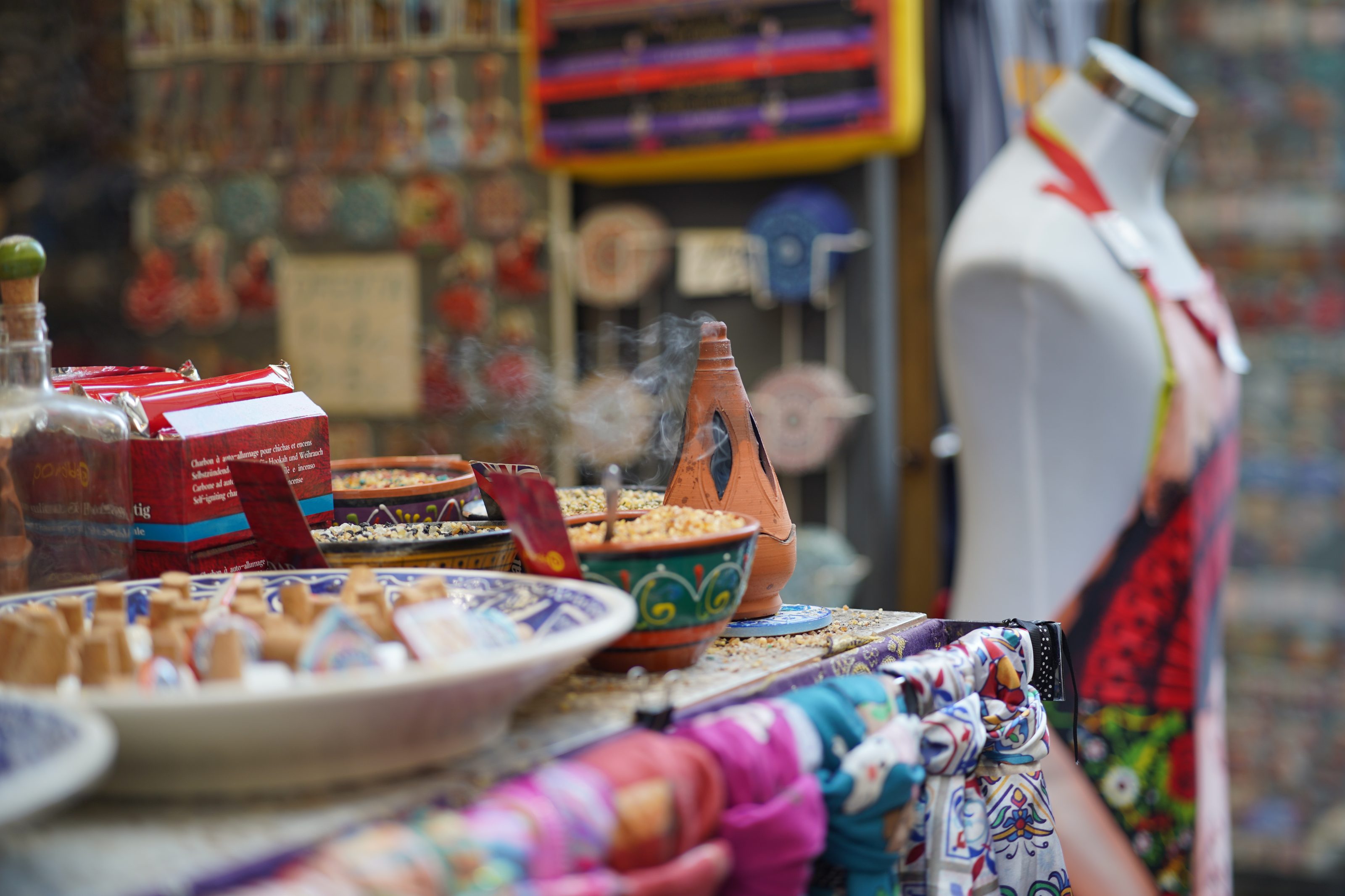 A market stall displaying colorful ceramic bowls filled with incense and spices, a terra cotta incense holder with smoke, vibrant scarves draped over the edge, and a mannequin wearing an apron in the background.