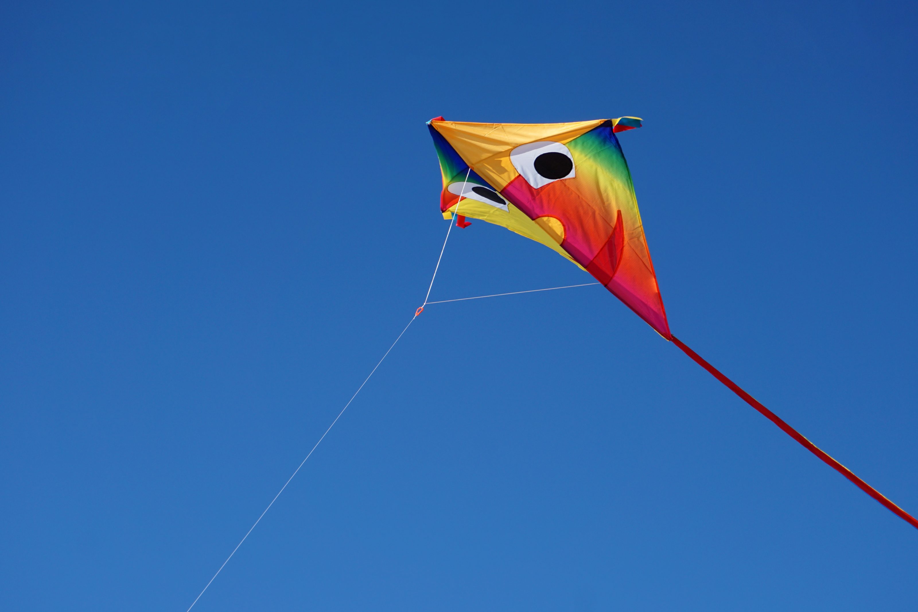 A colorful diamond-shaped kite with a face design, flying against a clear blue sky.