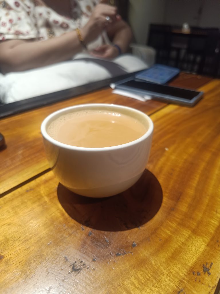 A white cup filled with tea on a wooden table. In the background, a person is holding a phone or small device, with another device lying on the table.