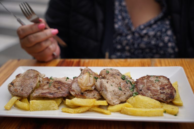 A plate of grilled meat and fried potatoes garnished with herbs (solomillo al whisky), with a person holding a fork and knife in the background.