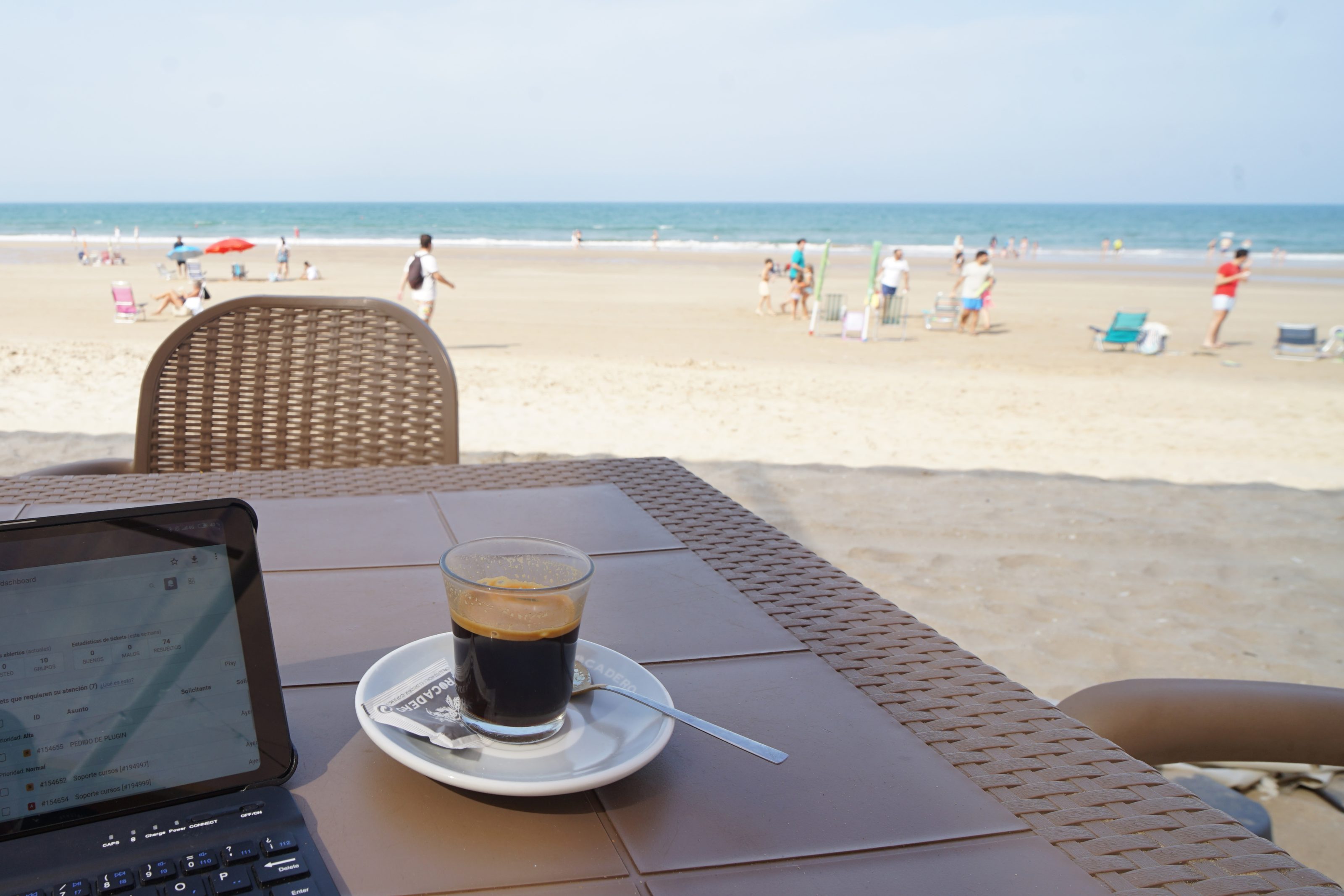 A beachside café table with a glass of coffee and a spoon, overlooking a sandy beach with people walking and relaxing near the ocean. A tablet is partially visible on the table.