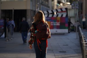 A person wearing a floral-patterned shirt and carrying a backpack is walking down a city street. The street is lined with metal barriers, and in the background, there is a large sign with colorful squares and a giant decorative clapperboard design.