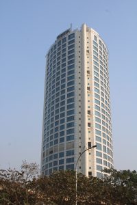 A tall, cylindrical skyscraper with numerous windows, set against a clear blue sky, with a streetlamp and foliage in the foreground.

