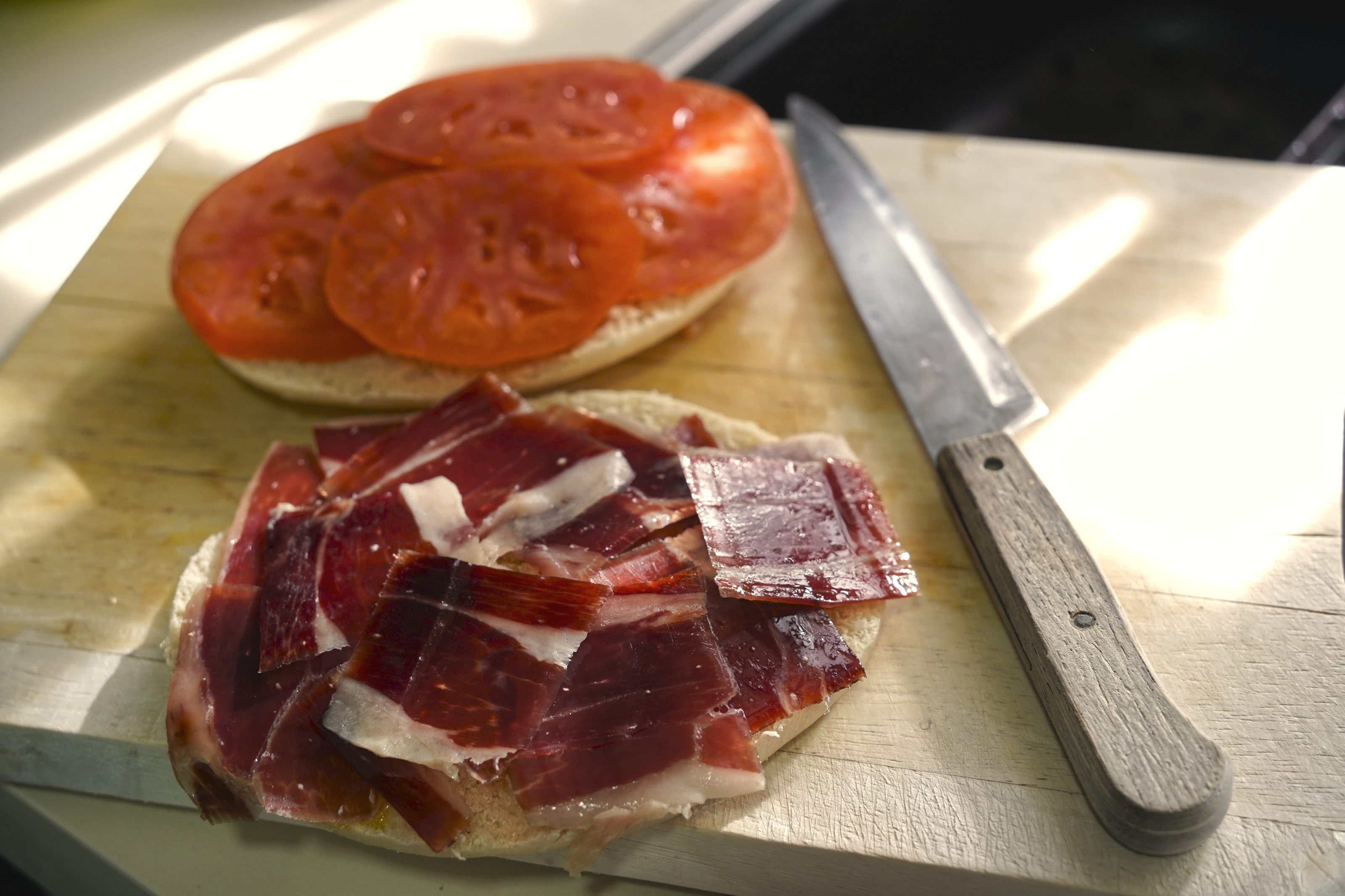 A sandwich being prepared on a wooden cutting board, with one half topped with slices of ibérico ham and the other half with slices of fresh tomato. A knife with a wooden handle is placed next to the sandwich.