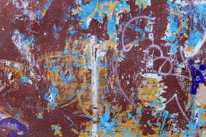 A textured wall with layers of peeling paint and graffiti. The background is primarily a rusty brown color with patches of vibrant blue, yellow, and white paint. Various graffiti markings and shapes are scattered across the surface, creating an abstract and weathered appearance.