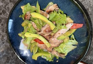 A vibrant salad served on a dark blue plate, featuring grilled chicken slices, avocado pieces, fresh lettuce, and tomato wedges