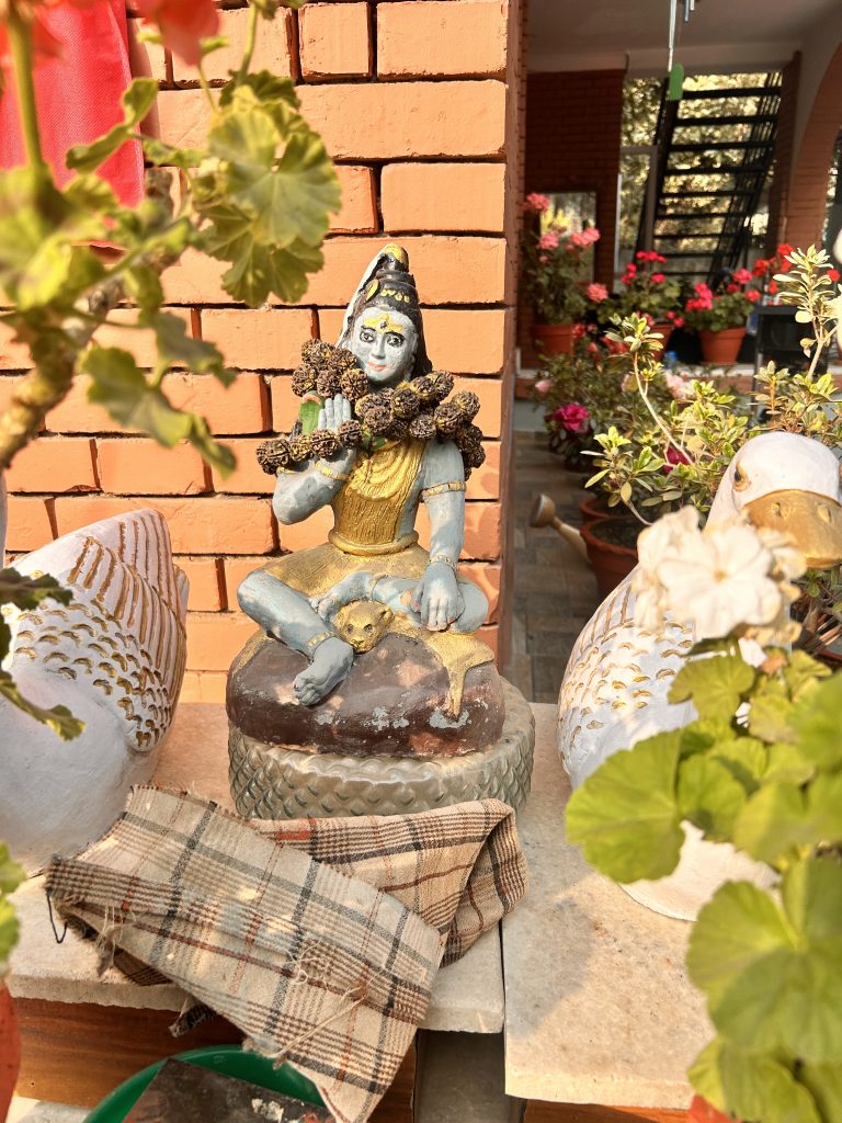 A small Shiva god statue of a blue-skinned deity adorned with a garland, sitting on a pedestal with a backdrop of brick wall.