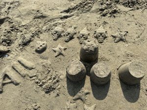 Fun sand art on the beach showing little alien faces and star shapes molded in the sand, with four cylindrical sand towers casting long shadows beneath them.