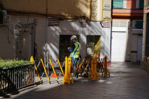 A construction worker wearing a safety vest and helmet is working on a street, surrounded by yellow barriers. The scene is set in a narrow alley with shop fronts and a sign on the wall.