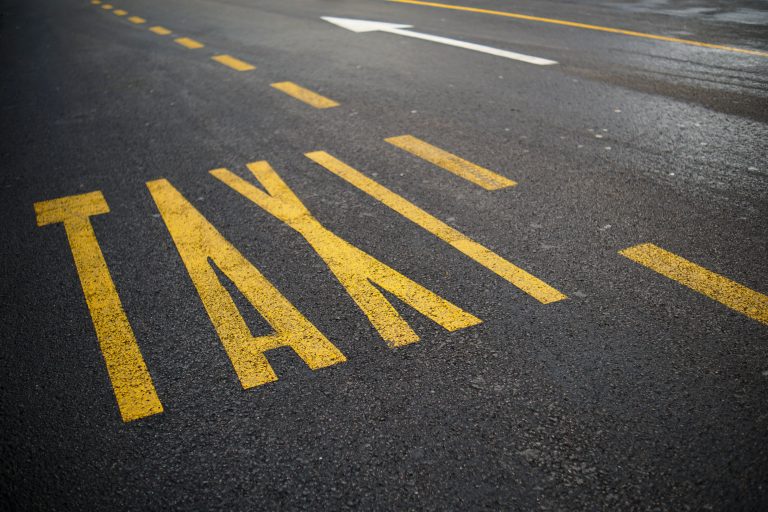 A road with the word “TAXI” painted in large yellow letters. There are dashed yellow lines and a white directional arrow on the asphalt.