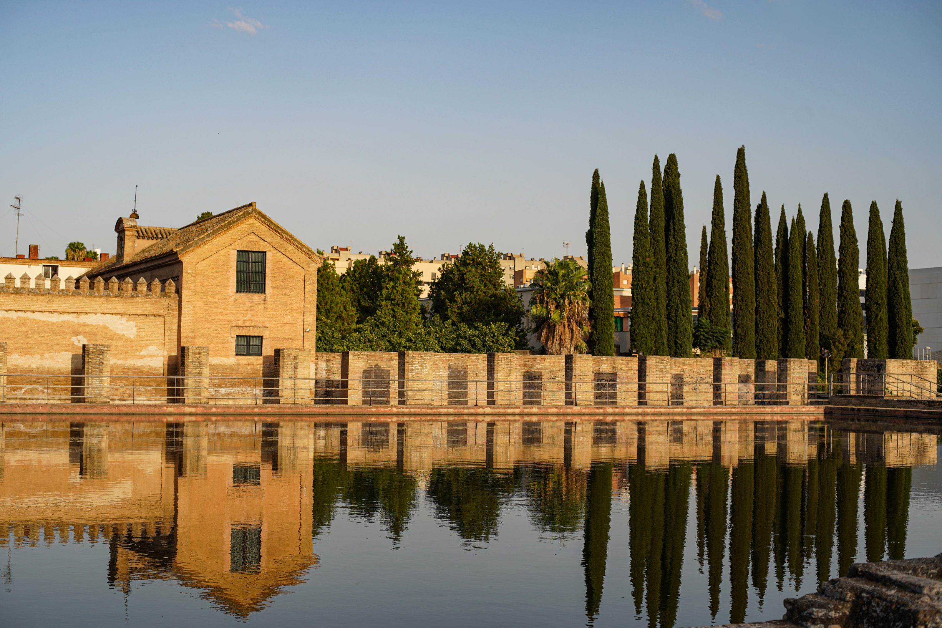 A historic brick building with a tile roof is reflected in a calm body of water. Tall cypress trees and a stone wall line the background, with a clear blue sky overhead.