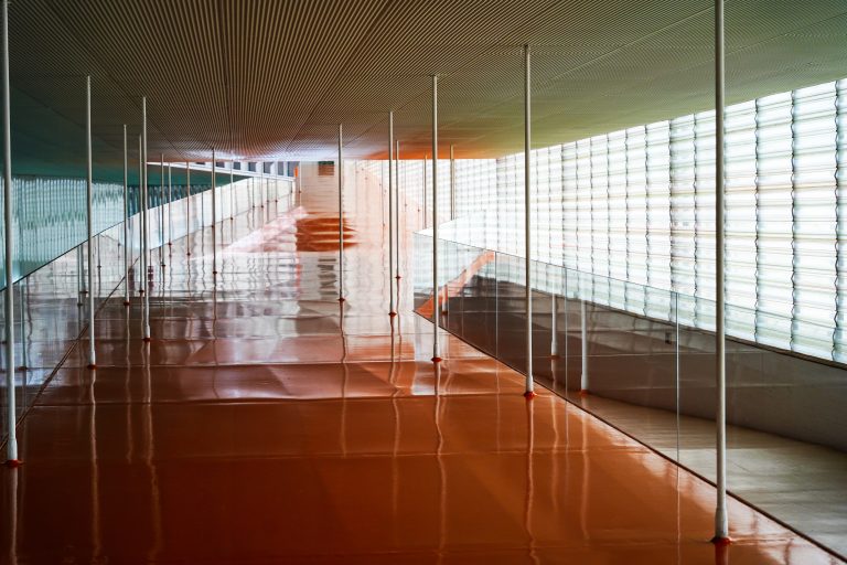 A modern, empty corridor with a shiny orange floor, translucent wall panels on the right, and a ceiling of parallel lines. Tall, thin support poles run alongside the pathway.