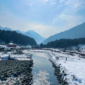 A snow-covered valley with a flowing river is surrounded by pine forests and mountains.