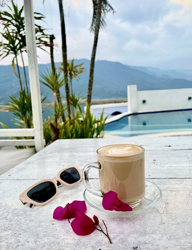 A cup of latte on a white wooden table with pink flowers and beige sunglasses, overlooking a pool, tropical plants, and a misty mountain view in the background.